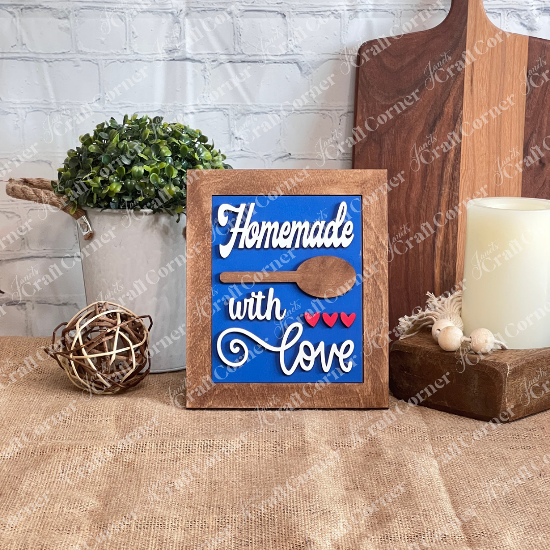 The Kitchen Switchable Farmhouse Sign by Janet's Craft Corner features a blue wooden sign with "Homemade with love" in white, a spoon cutout, and red hearts—ideal rustic decor for your kitchen. Set of 1, perfect for DIY home decorating.
