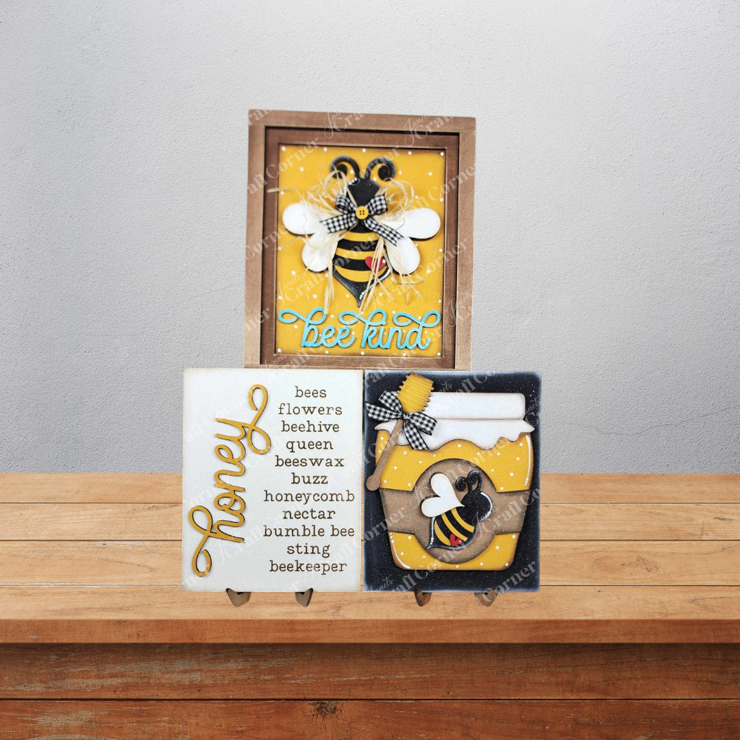 Samples of the Honey Bee Farmhouse Signs by Janet’s Craft Corner. Each is made from a delightful DIY home décor craft kit featuring three options: one that says “bee kind” with a buzzing bee, one with bee-related words, and one with a honey jar design.