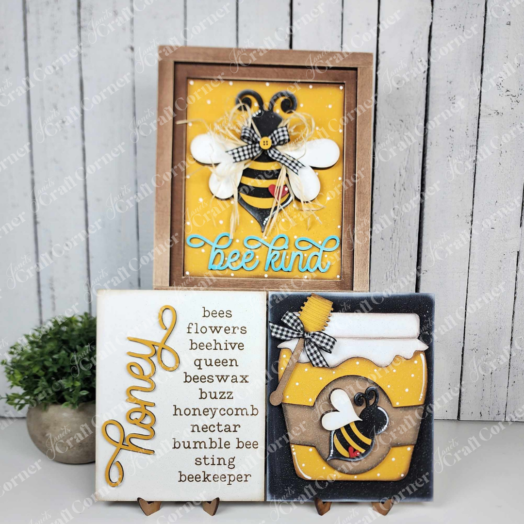 Samples of the Honey Bee Farmhouse Signs by Janet’s Craft Corner. Each is made from a delightful DIY home décor craft kit featuring three options: one that says “bee kind” with a buzzing bee, one with bee-related words, and one with a honey jar design.