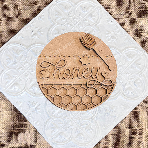 The Honey Bee Mini Shelf Sitters by Janet's Craft Corner is a DIY decor kit featuring a round wooden plaque with "honey" in cursive, bee and heart accents, a honey dipper, and honeycomb design—perfect for seasonal home decorating.