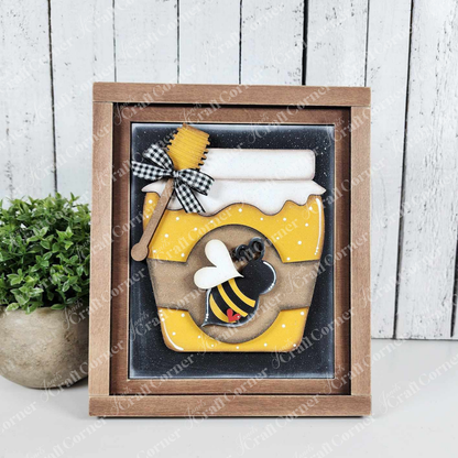 This sample of the Honey Jar Farmhouse Sign from Janet's Craft Corner features a charming black-and-white bow and bee design with a honey dipper on top, set against a weathered white wood background with a small green plant, ideal for farmhouse decor lovers.