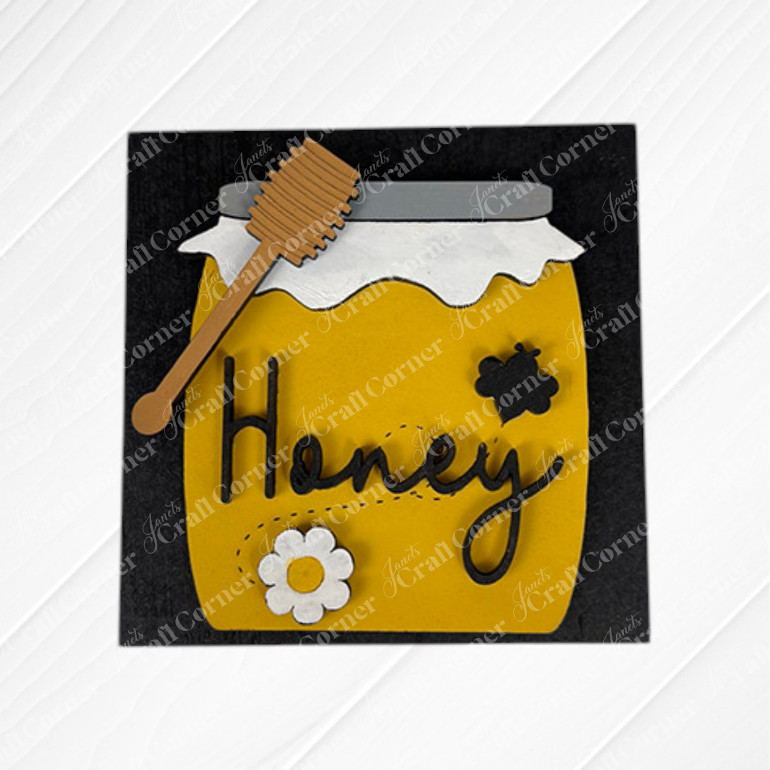 The Bee Swappable Squares from Janet's Craft Corner feature a yellow honey jar with a white lid, wooden dipper, white flower, and black bee on customizable squares. "Honey" is scripted in cursive on a black background for unique decor.