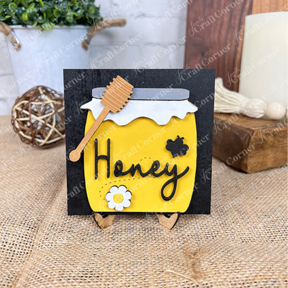 Janet's Craft Corner Bee Swappable Squares kit features a handmade card with a yellow honey jar, "Honey" label, wooden dipper, flower and bee cutouts, and displays upright on a small wooden stand for charming décor.