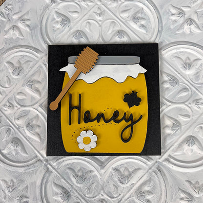 The Bee Swappable Squares from Janet's Craft Corner feature a yellow honey jar with a white lid, wooden dipper, white flower, and black bee on customizable squares. "Honey" is scripted in cursive on a black background for unique decor.