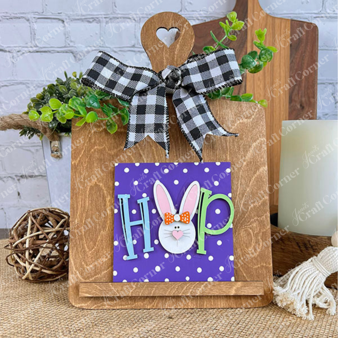 Janet's Craft Corner’s Easter Bunny Squares features a "HOP" sign with a bunny face on a purple polka-dot background, displayed on a wooden easel with a checkered bow and greenery—perfect for your tiered tray decor.