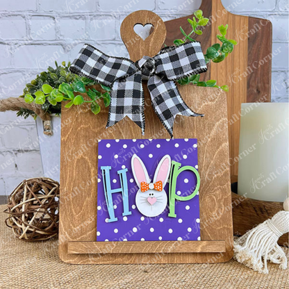Janet's Craft Corner’s Easter Bunny Squares features a "HOP" sign with a bunny face on a purple polka-dot background, displayed on a wooden easel with a checkered bow and greenery—perfect for your tiered tray decor.