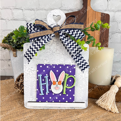 Janet's Craft Corner Easter Bunny Squares: A white wooden board with a heart cutout, green leaves, and a chevron bow, paired with a purple polka-dot Swappable Square Display that spells “HOP” with a bunny face for the "O". Perfect for tiered tray decor.