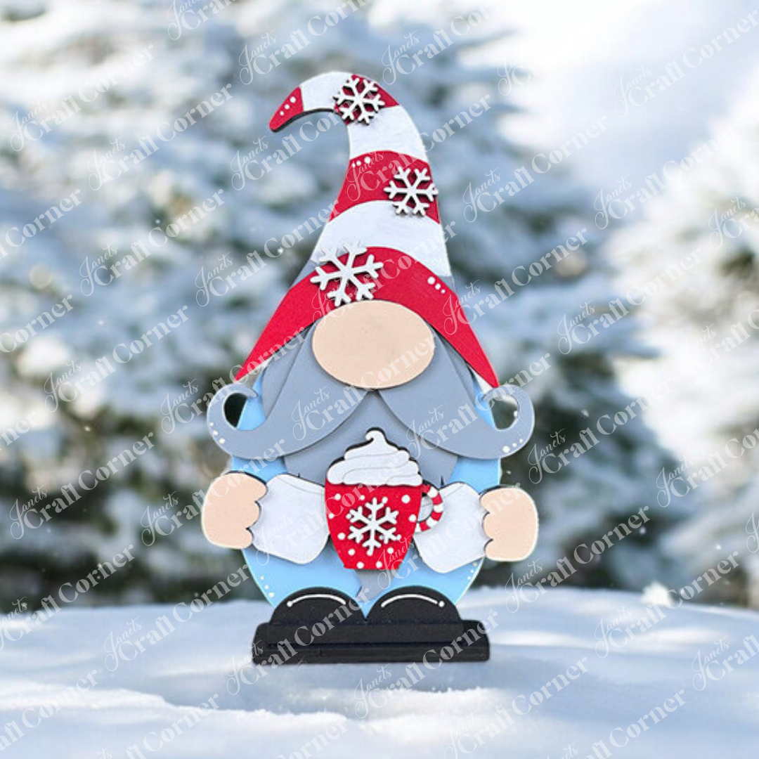 A Hot Cocoa Gnome Shelf Sitter from Janet's Craft Corner stands with a red and white snowflake hat, holding a matching mug in a snowy landscape. Ideal for holiday decor, blurred snow-covered trees complete this cozy winter scene.