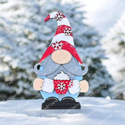 A Hot Cocoa Gnome Shelf Sitter from Janet's Craft Corner stands with a red and white snowflake hat, holding a matching mug in a snowy landscape. Ideal for holiday decor, blurred snow-covered trees complete this cozy winter scene.