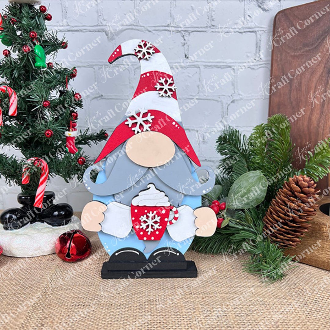 The Hot Cocoa Gnome from Janet's Craft Corner is a charming festive figurine holding a red mug of steaming hot cocoa, complete with a swirl of whipped cream. This delightful gnome features a tall hat adorned with snowflakes and is accompanied by a small decorated Christmas tree, ornaments, and pine cones on a rustic burlap surface—ideal for adding charm to your home decor.