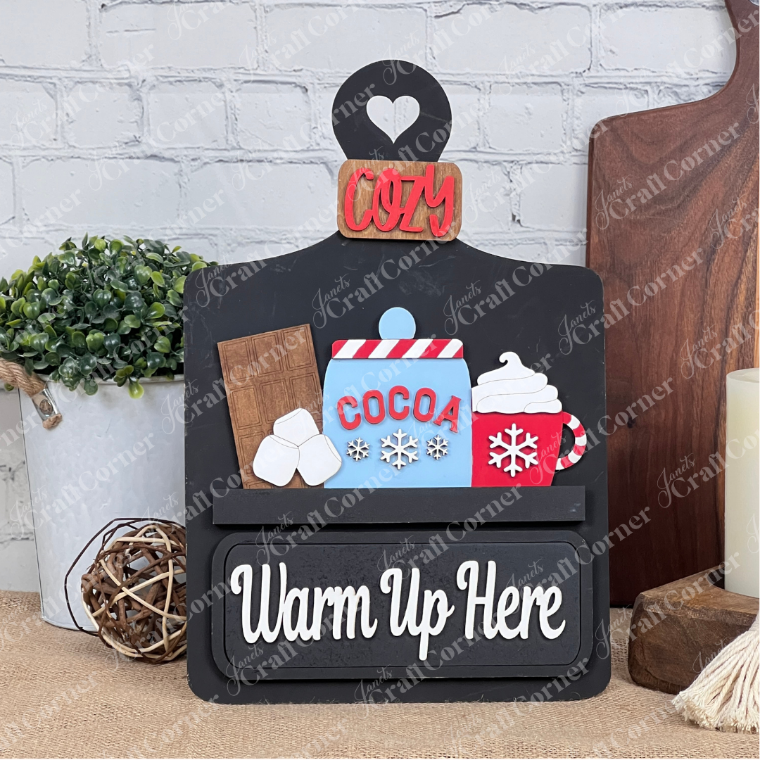 Janet's Craft Corner presents a DIY home decor kit featuring a "Warm Up Here" sign with cocoa illustrations. The included insert has decorative elements like whipped cream, marshmallows, a red "Cozy" label, and background details resembling a plant and candle on wooden boards.