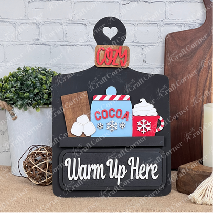 Janet's Craft Corner presents a DIY home decor kit featuring a "Warm Up Here" sign with cocoa illustrations. The included insert has decorative elements like whipped cream, marshmallows, a red "Cozy" label, and background details resembling a plant and candle on wooden boards.