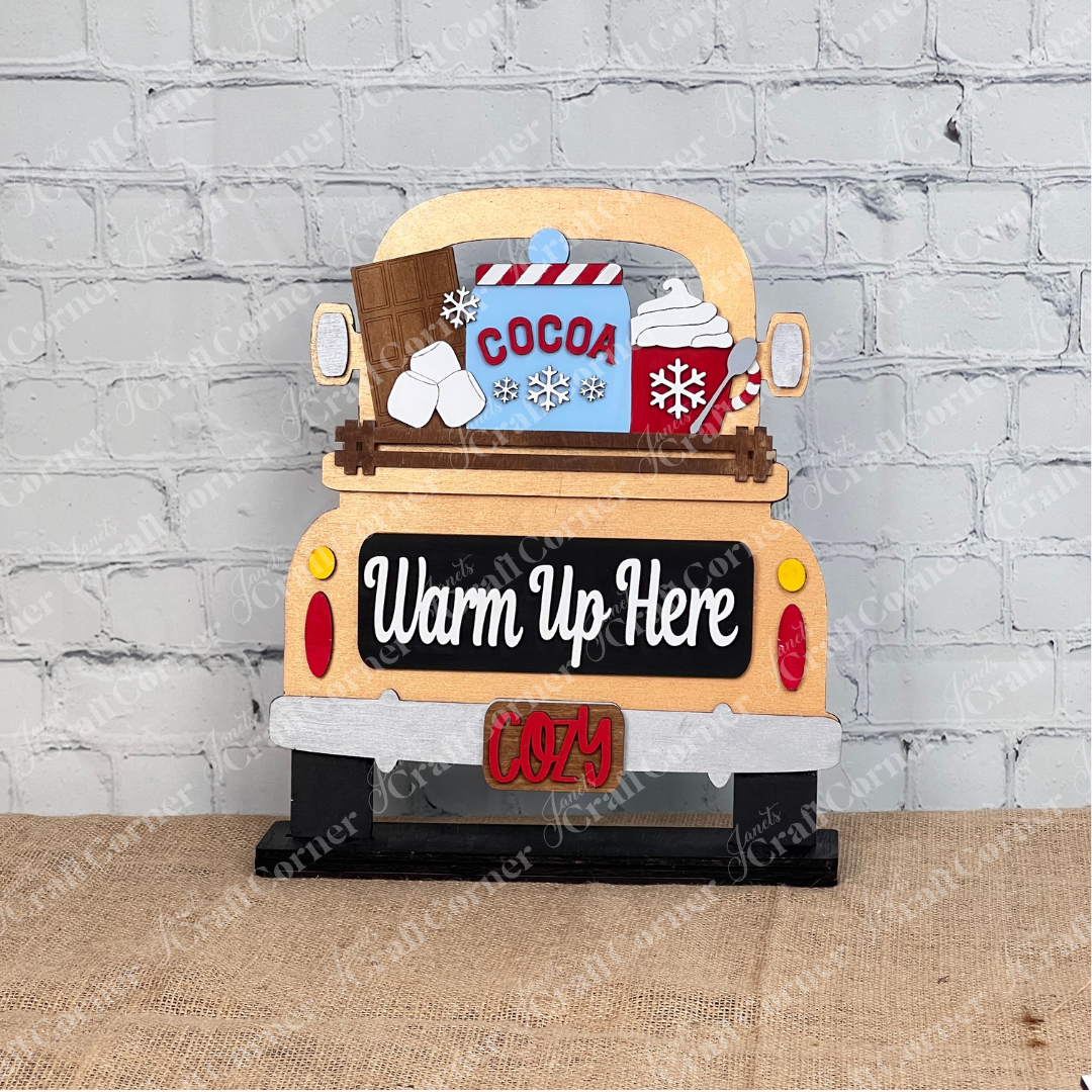 The Hot Cocoa Interchangeable Insert by Janet's Craft Corner is a DIY seasonal home decor craft kit featuring a truck bed with a "Cocoa" mug, whipped cream, snowflakes, and logs. The bumper reads "Cozy," all set against a white brick wall on burlap, perfect for creative customization.