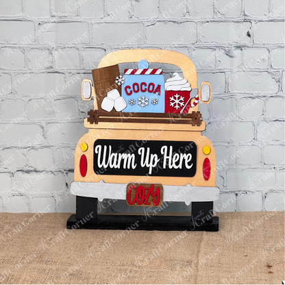 The Hot Cocoa Interchangeable Insert by Janet's Craft Corner is a DIY seasonal home decor craft kit featuring a truck bed with a "Cocoa" mug, whipped cream, snowflakes, and logs. The bumper reads "Cozy," all set against a white brick wall on burlap, perfect for creative customization.