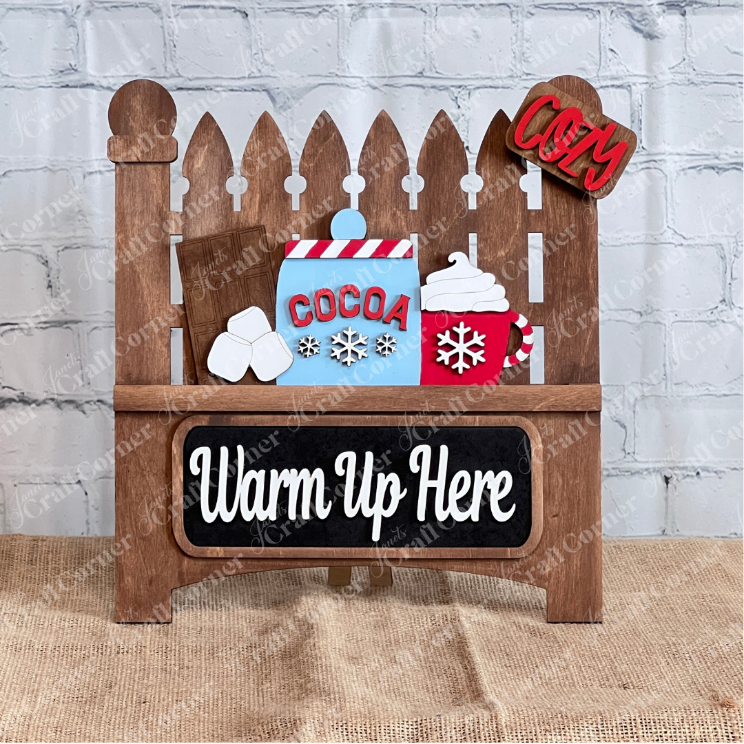 The Janet's Craft Corner "Garden Fence with Insert" is a DIY home decor kit featuring a decorative wooden sign with hot cocoa elements—marshmallows, a chocolate bar, and a steaming cup with snowflakes. The message "Cocoa Warm Up Here" evokes cozy vibes against its brick wall and burlap backdrop.