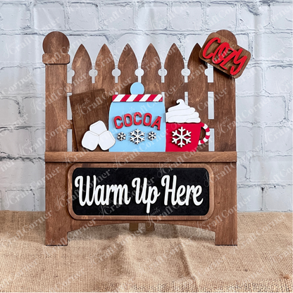 The Janet's Craft Corner "Garden Fence with Insert" is a DIY home decor kit featuring a decorative wooden sign with hot cocoa elements—marshmallows, a chocolate bar, and a steaming cup with snowflakes. The message "Cocoa Warm Up Here" evokes cozy vibes against its brick wall and burlap backdrop.