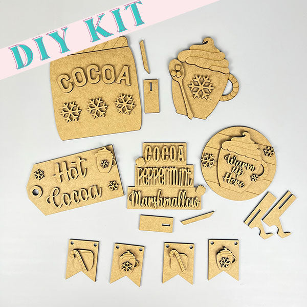 Flat lay of Janet's Craft Corner's Hot Cocoa Tiered Tray Pieces, a holiday décor DIY kit for a hot cocoa-themed craft project. The kit includes cutouts of a cocoa mug, marshmallows, snowflakes, and decorative tags with phrases like "Hot Cocoa" and "Warm Here," perfect for creating your own tiered tray with wooden pieces and string.