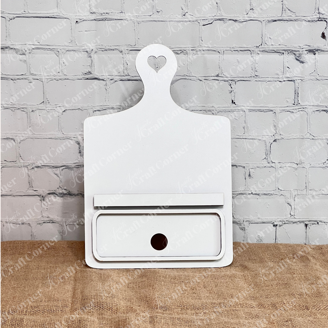Janet's Craft Corner offers the Breadboard Classic Interchangeable Base with Insert DIY kit. This decorative white board has a heart handle, minimalistic face markings, and interchangeable accessories—ideal for DIY enthusiasts against a burlap backdrop.