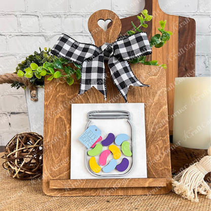 Hello Easter Squares by Janet's Craft Corner is a wooden cutting board with a heart cutout, adorned with a plaid bow and greenery. Featuring a coffee-themed jar of colorful jellybeans, it makes the perfect decorative accent.
