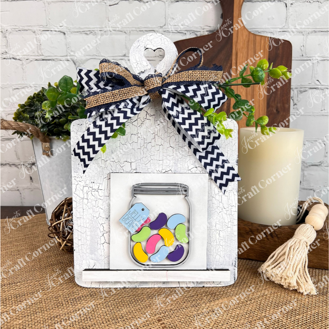 Janet's Craft Corner's "Hello Easter Squares" is a white rustic plaque with a heart cutout, chevron bow, greenery, and a jelly bean jar—perfect for farmhouse or coffee-themed decor alongside candles and cozy accents.