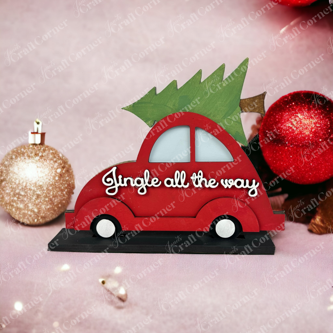 The "Jingle All the Way Volkswagen Shelf Sitter" by Janet's Craft Corner is a decorative red figurine with an evergreen tree on top, surrounded by red and gold Christmas baubles against a soft pink background with twinkling lights, adding yuletide cheer.