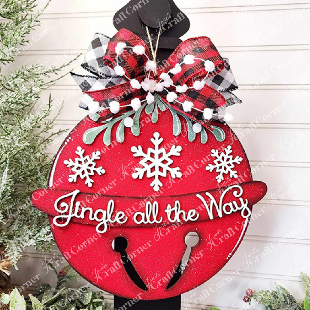 The Jingle Bell Door Hanger by Janet's Craft Corner adds a festive touch to your holiday decor with its red ornament adorned with white snowflakes and the phrase "Jingle all the Way" in cursive. It is completed with a black and white plaid bow, green foliage accents, and a backdrop of snow-dusted greenery.