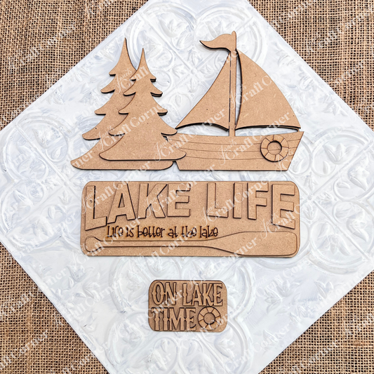 Janet's Craft Corner Lake Life Interchangeable Insert is a DIY craft kit featuring 3 wooden pieces—two pine trees, a sailboat, and two signs with "Lake Life" phrases—perfect for seasonal home decor.