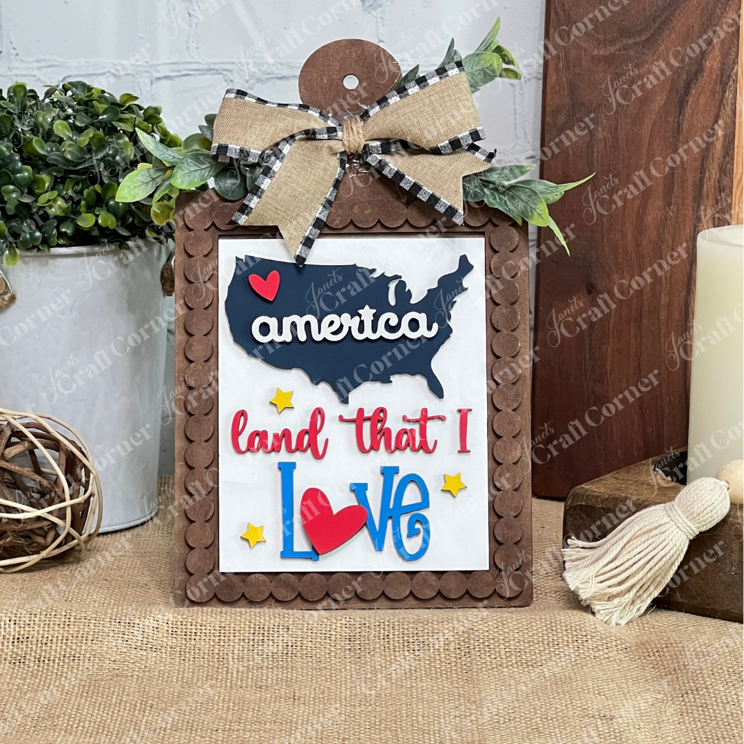 The Patriotic Farmhouse Sign DIY craft kit by Janet's Craft Corner features a wooden-framed USA map with "America land that I love," heart, stars, plaid bow, and greenery—perfect tiered tray decor among plants, candles, and tassels.