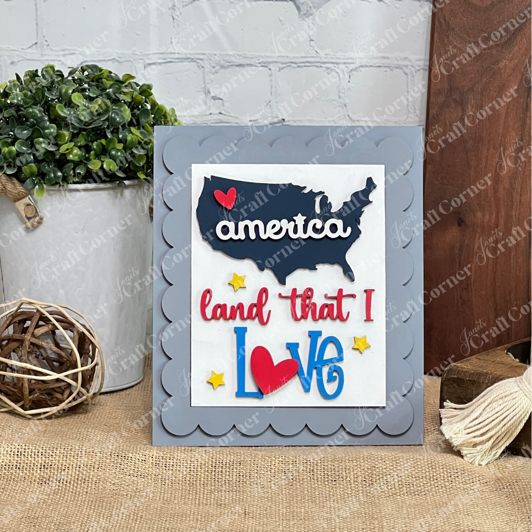 Janet's Craft Corner Patriotic Farmhouse Sign is a DIY decor kit featuring a U.S. map cutout with "America" in white script, a red heart, “land that I love” in red and blue, yellow stars, and a scalloped gray frame. Set of 1.