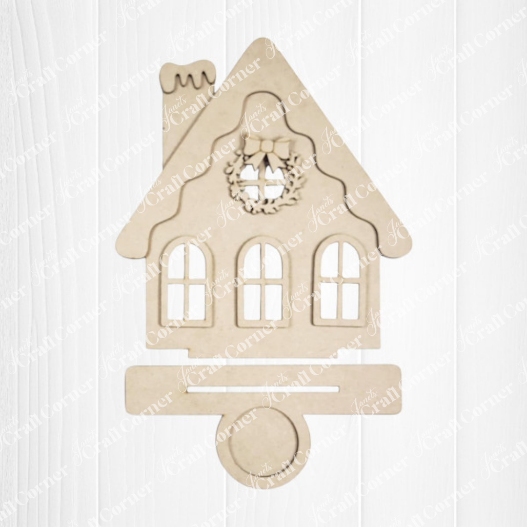 The Victorian Gingerbread House Votive Holder DIY craft kit by Janet's Craft Corner features a festive wooden cutout with a wreath above the windows and a slot at the bottom, ideal for seasonal decor, displayed on a white wood background.