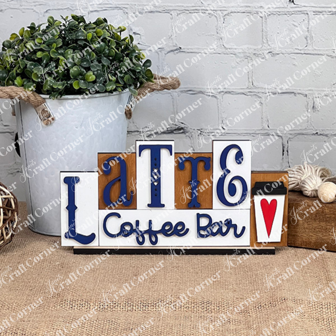 This charming home décor piece from Janet's Craft Corner, called the Latte Mini Word Block, displays "Latte Coffee Bar" in blue and black on a white background. It includes a delightful coffee cup illustration with a red heart, making it perfect for coffee lovers and adding warmth to any space when displayed against its white brick wall setting.