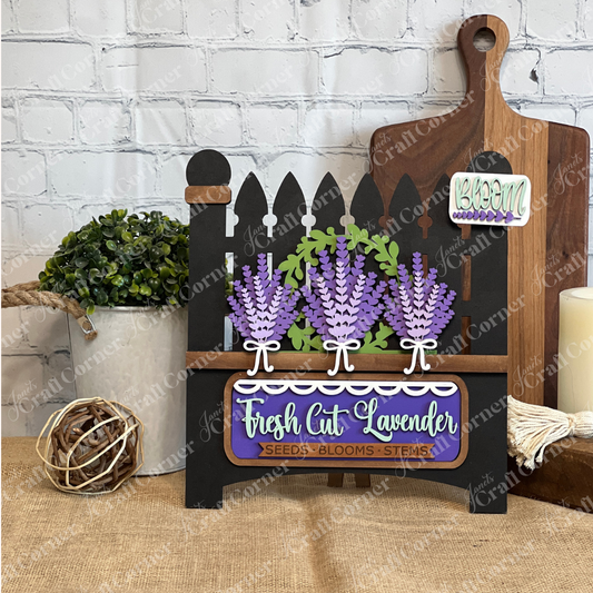 The Lavender Interchangeable Insert DIY Craft Kit by Janet's Craft Corner features a black picket fence sign with purple lavender, “Fresh Cut Lavender” and “Bloom” labels—ideal farmhouse décor for tables with potted plants or cutting boards.