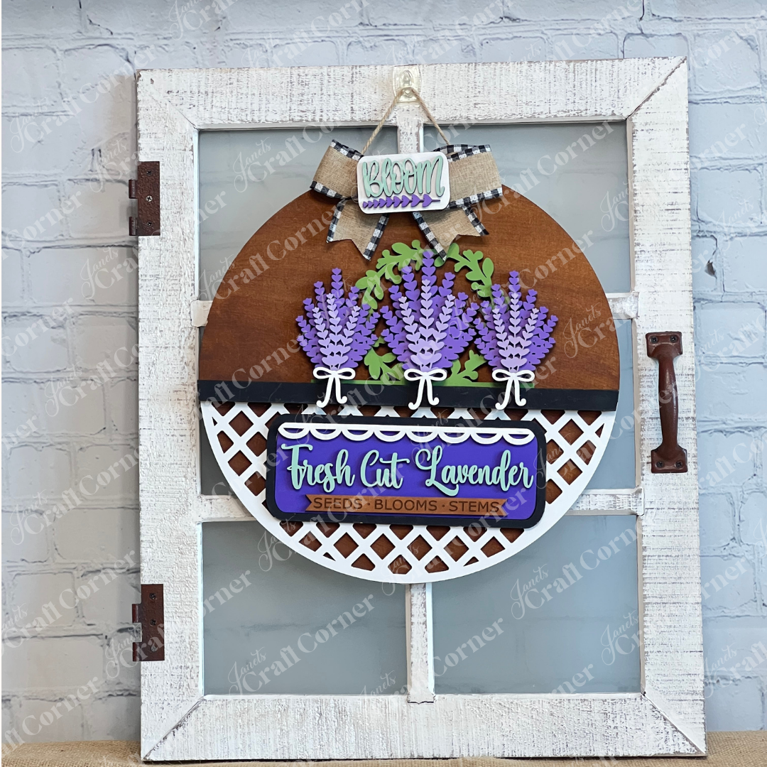 The Lavender Interchangeable Insert DIY Craft Kit from Janet's Craft Corner features purple lavender and green leaves with a "Bloom" sign and bow, perfect for farmhouse or lavender-themed décor against a white brick wall.