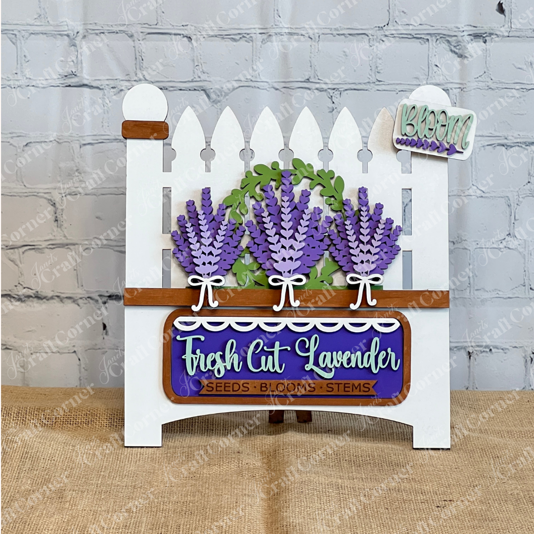 Elevate your home décor with the Lavender Interchangeable Insert DIY Craft Kit from Janet's Craft Corner, featuring a white picket fence, three purple lavender bunches, leafy greenery, and farmhouse signs—perfect for a brick wall accent.