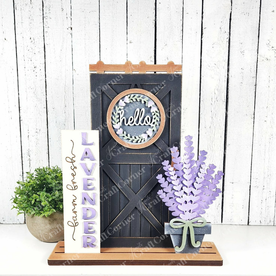 Create a charming rustic look with Janet's Craft Corner Lavender Interchangeable Porch Set, featuring a black "hello" barn door sign, vertical unfinished wood "LAVENDER" sign, faux lavender in a gray pot, and greenery on a white wooden backdrop.