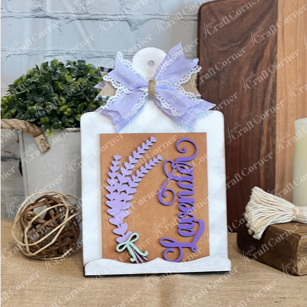 Janet's Craft Corner Lavender Switchable Farmhouse Signs DIY Craft Kit features a decorative sign with lavender cutouts, purple "Lavender" script, and a bow—ideal tiered tray décor. Displayed with rustic accents and other switchable signs.