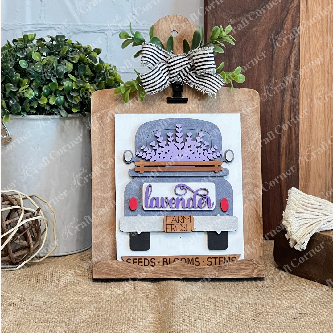 The Lavender Switchable Farmhouse Signs – DIY Craft Kit by Janet's Craft Corner features a vintage truck with lavender flowers, clipped to a wooden board with a striped bow, perfect for adding rustic charm to your tiered tray décor.