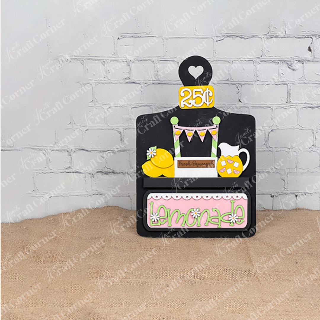 Welcome summer with Janet's Craft Corner's Lemonade Interchangeable Insert, a charming 3-piece DIY home decor kit featuring a chalkboard-style sign with a heart-shaped "25¢," lemonade pitcher, and lemon illustrations. "Fresh Squeezed Lemonade" enhances the design amid floral accents on brick and burlap.