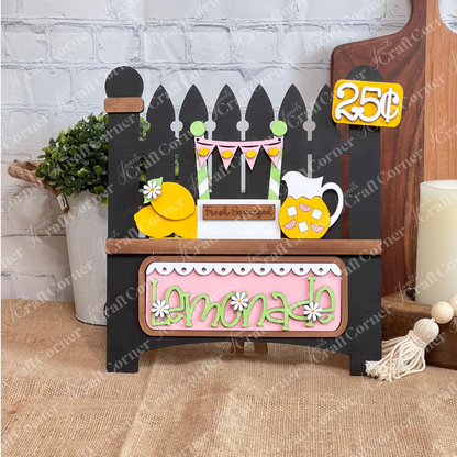 Janet's Craft Corner offers a DIY seasonal home decor craft kit called the Lemonade Interchangeable Insert. It features a charming black picket fence design with lemons, an ice-filled pitcher, a "Fresh Squeezed" label, and an interchangeable pink insert with "Lemonade," elegantly displayed in white and green letters.