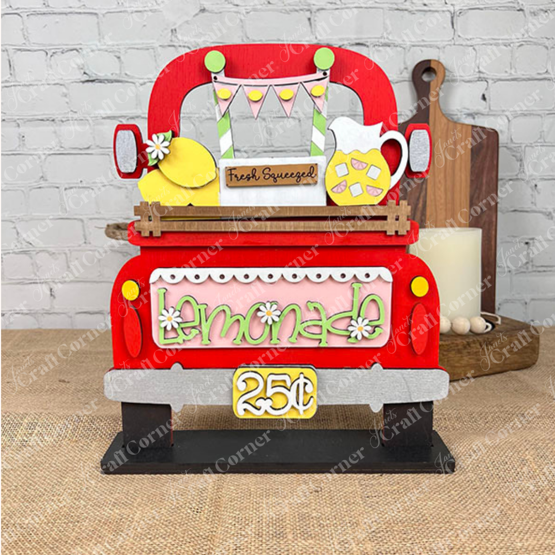 The Lemonade Interchangeable Insert DIY home decor kit by Janet's Craft Corner includes a charming hand-painted wooden display featuring a red truck with a "lemonade" sign priced at 25 cents. It comes complete with a lemon, pitcher, and "Fresh Squeezed" banner set against a brick wall backdrop, making it an ideal choice for home décor or as an interchangeable insert.