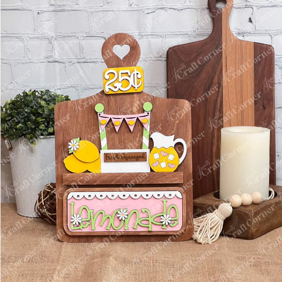 Welcome summer with Janet's Craft Corner's Lemonade Interchangeable Insert, a charming 3-piece DIY home decor kit featuring a chalkboard-style sign with a heart-shaped "25¢," lemonade pitcher, and lemon illustrations. "Fresh Squeezed Lemonade" enhances the design amid floral accents on brick and burlap.