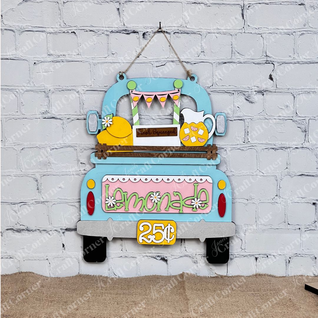 The Lemonade Interchangeable Insert from Janet's Craft Corner features a blue truck with lemons, pink "Lemonade" letters, and a "Fresh Squeezed" sign. A "25¢" price tag adds nostalgia to this DIY home decor craft kit, perfect for hanging on a brick wall.