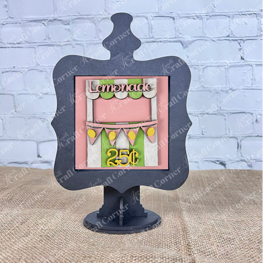 The Lemon Squares sign by Janet's Craft Corner features a lemonade stand illustration with green and pink accents, lemon slices, and "Lemonade 25¢" text. It sits on a black stand against burlap and a white brick wall.