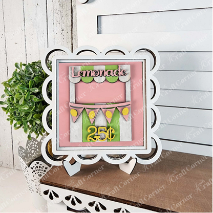 Janet's Craft Corner's "Lemon Squares" decorative framed sign on a stand displays a pink and green lemonade stand design with yellow bunting and "Lemonade" and "25¢" text, perfect for DIY seasonal home decor beside a plant.