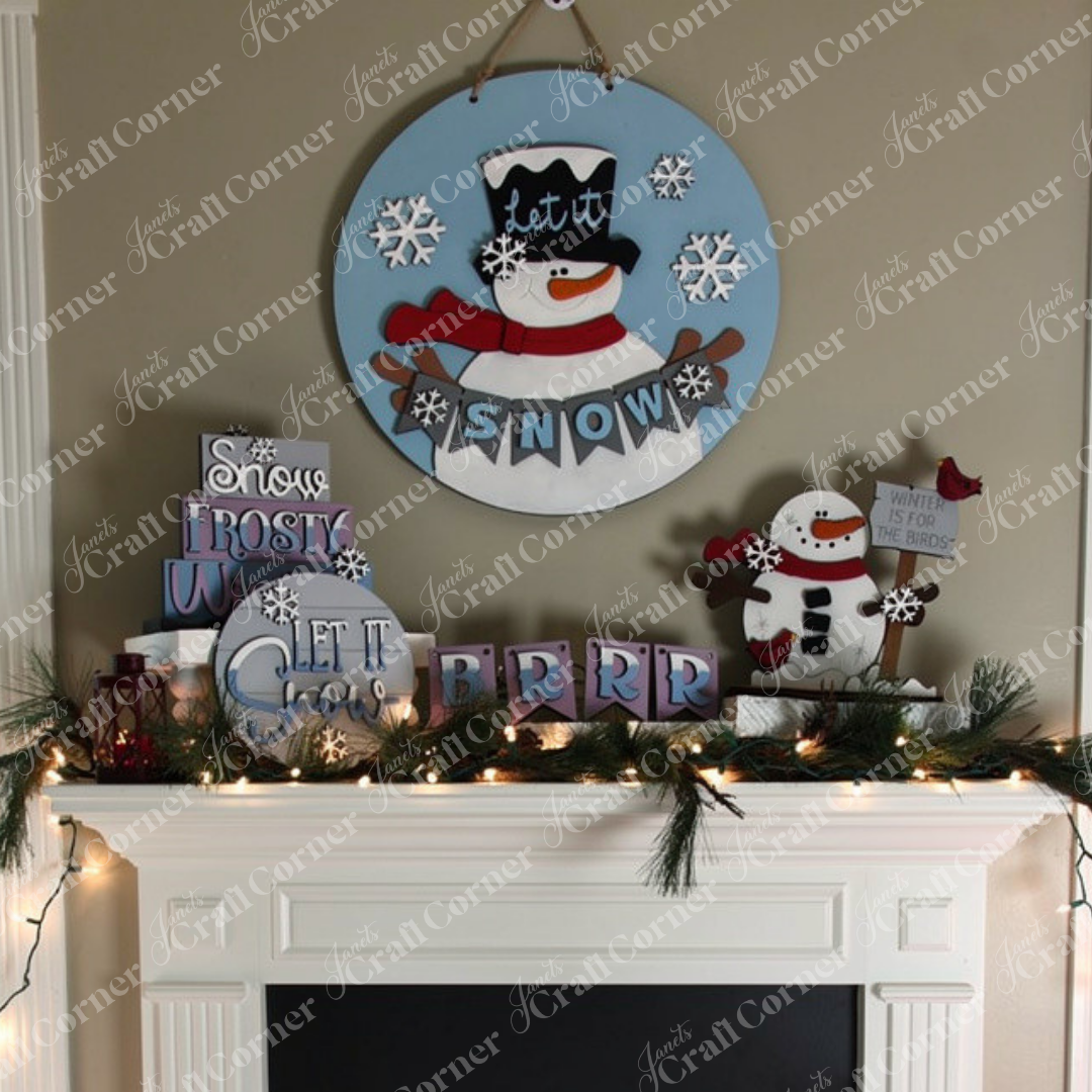 A festive winter display featuring Janet's Craft Corner's "Let It Snow Tiered Tray" showcases a snowman design wall hanging adorned with "Let it Snow" text, surrounded by charming holiday decor. The display includes signs with messages like "Snow," "Frosty Winter," "Let It Snow," and "BRRR," displayed on and above a mantle with white mini lights and a greenery garland for a magical touch.