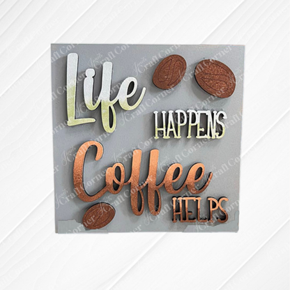The Coffee Swappable Squares tabletop sign from Janet's Craft Corner features green and brown raised letters on a gray background reading "Life happens, coffee helps," displayed on a white wood background.