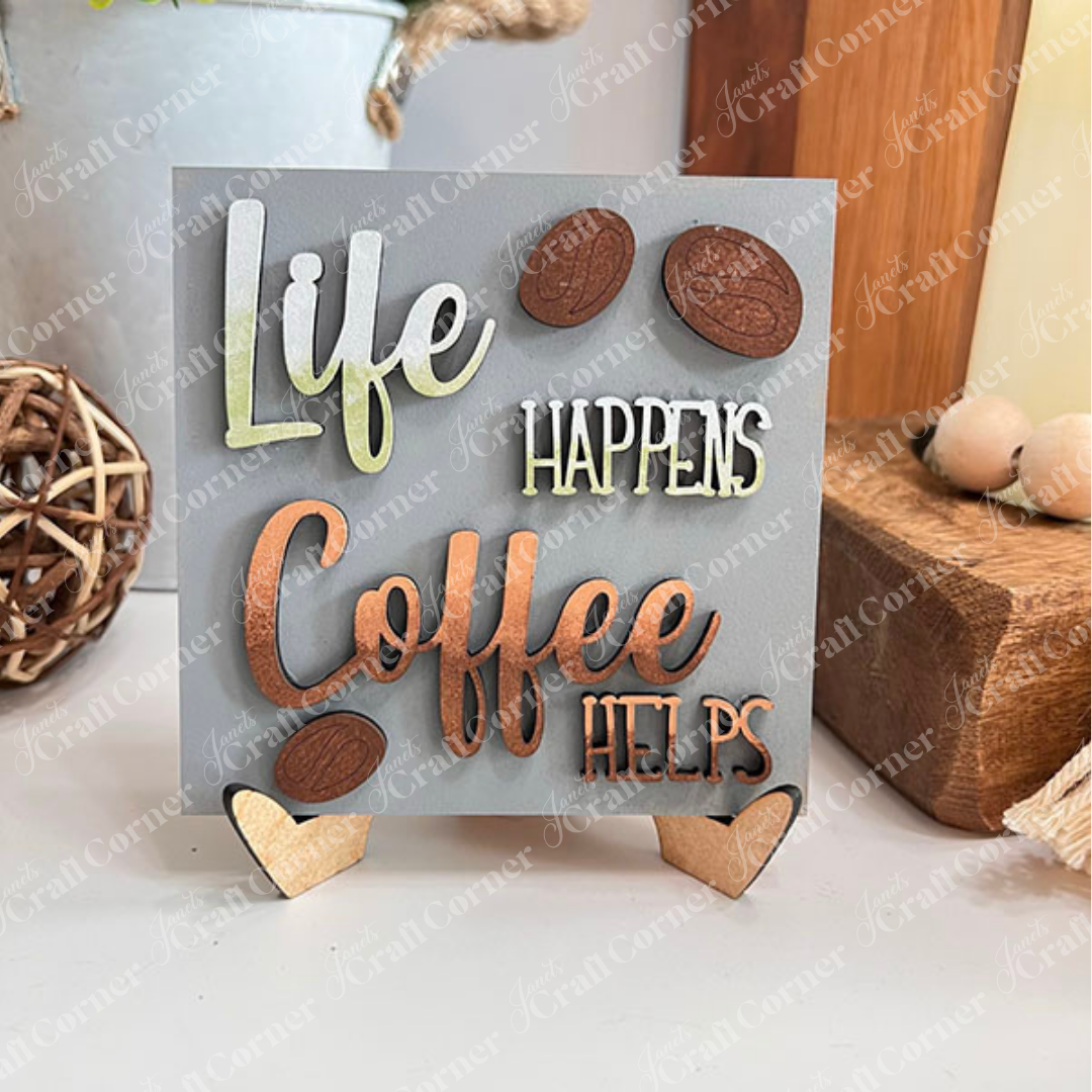 The Coffee Swappable Squares tabletop sign from Janet's Craft Corner features green and brown raised letters on a gray background reading "Life happens, coffee helps," making it an ideal DIY home decor accent for any space.