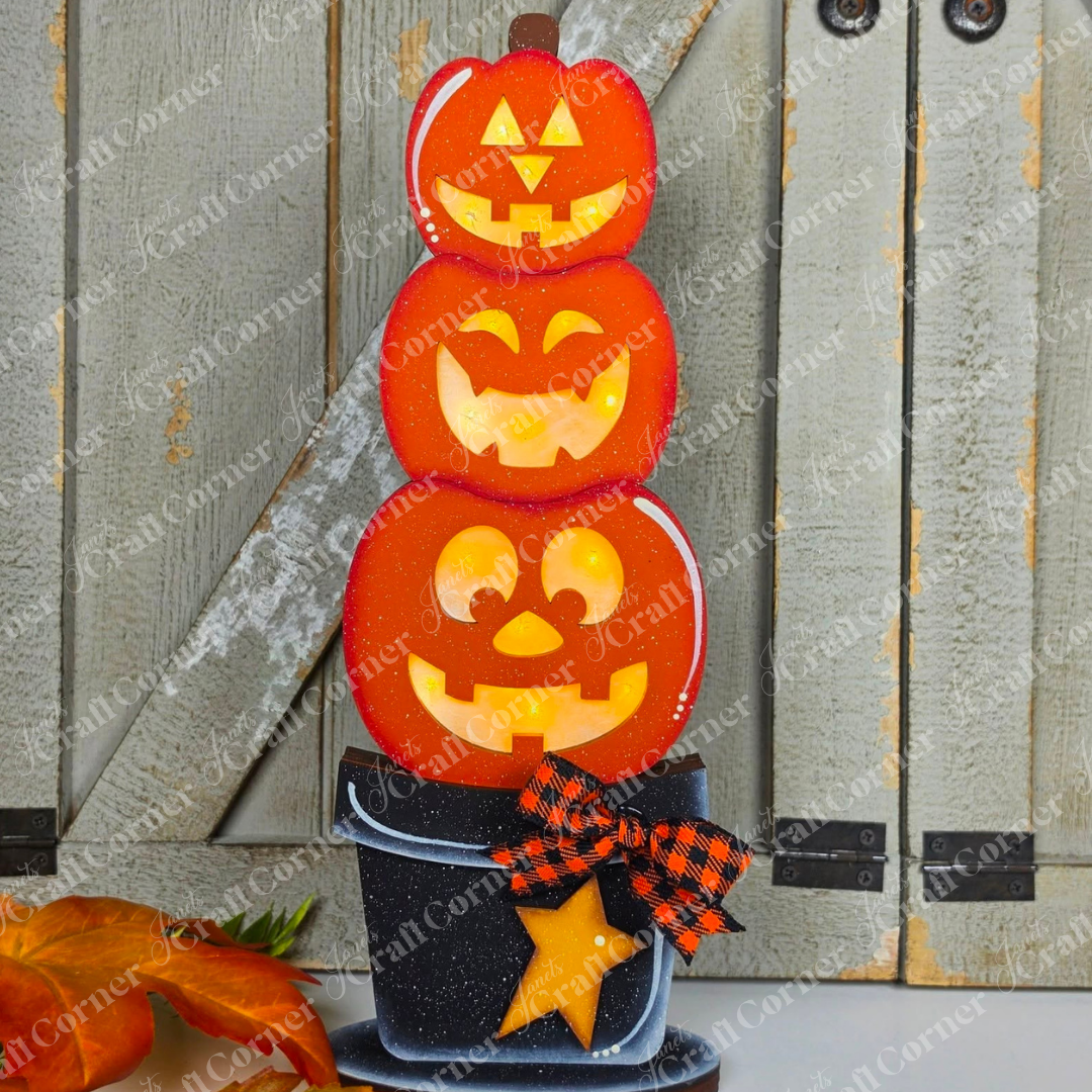 Janet’s Craft Corner Light-up Stacking Pumpkin Shelf Sitter, available as a DIY home decor craft kit in your choice of two sizes, features three stacked jack-o’-lanterns with glowing faces sitting in a black pot decorated with an orange star and bow. Displayed against wooden barn doors and surrounded by autumn leaves, this charming shelf sitter is the perfect addition to your fall and Halloween decor.