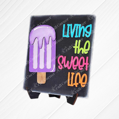 Janet's Craft Corner Summer Switchable Farmhouse Sign, a DIY kit with a purple popsicle design and "Living the sweet life" text, adds rustic charm to any space. Set of 1 includes mini easel for display.