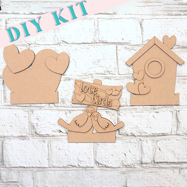 The Love Birds Train Inserts DIY craft kit from Janet's Craft Corner includes cardboard cutouts of two charming lovebirds with a "love birds" sign, a birdhouse, and cloud shapes adorned with hearts. Ideal for Valentine's inserts, it features a white brick background.